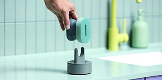 Ein Brite by Scotch-Brite™ Spülschwamm mit Griff wird auf einen Schwammhalter auf der Küchenarbeitsplatte neben der Spüle gelegt.

